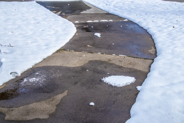 Close-up of curving residential sidewalk cleared of snow, but with spots remaining and large wet areas. Frost Heave